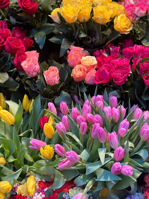 A vibrant floral display featuring a variety of fresh flowers including pink and yellow tulips in the foreground with smooth, green leaves, and a mix of pink, orange, and yellow roses arranged in clus
