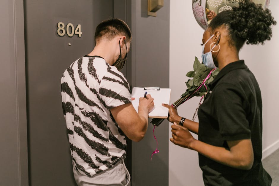 A man with short hair and a patterned black and white t-shirt is standing in front of a grey door numbered 804, writing on a clipboard held by a woman with curly hair, wearing a black top and face mas