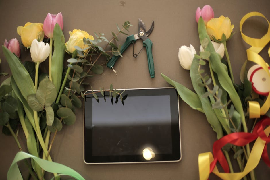 A flat-lay arrangement of fresh pastel-colored tulips and yellow roses with green foliage, placed on a neutral surface. The bouquet is wrapped with colorful ribbons—yellow, red, and green—adding d
