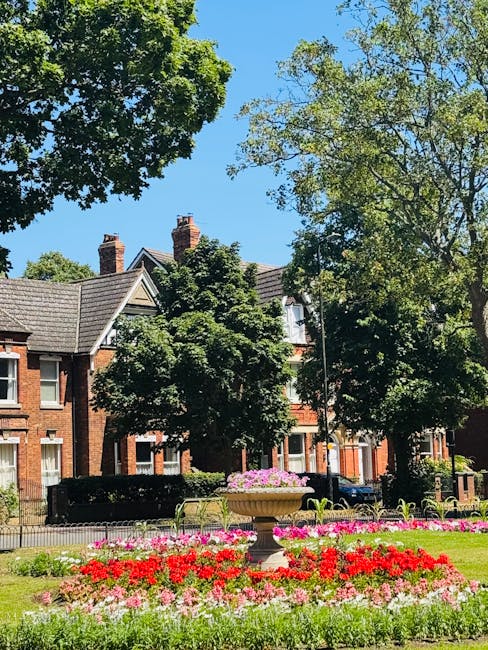 A vibrant garden scene featuring an array of colorful flowers arranged in neatly maintained flower beds with red, pink, and white blooms. In the center, there is a large stone or concrete planter fill