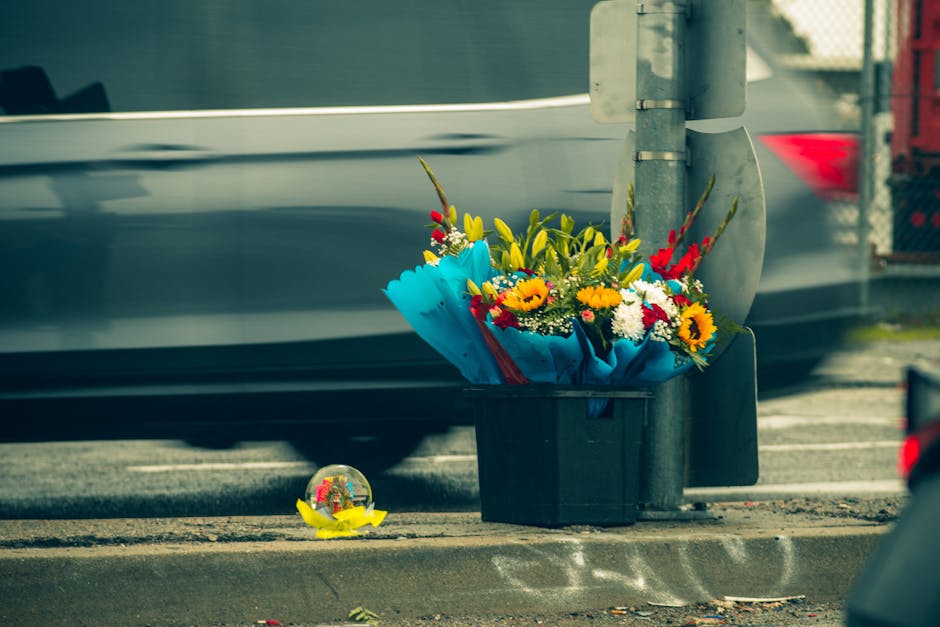 A vibrant floral bouquet placed in a black plastic container on a sidewalk next to a metal pole, featuring a mix of yellow daisies, red gladiolus, white chrysanthemums, and blue wrapping paper, with f