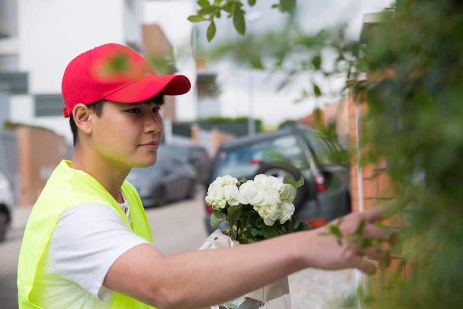A young male florist wearing a red cap and a neon yellow vest is carefully arranging a bouquet of fresh white hydrangeas with green leaves, holding the flowers with one hand while adjusting them with